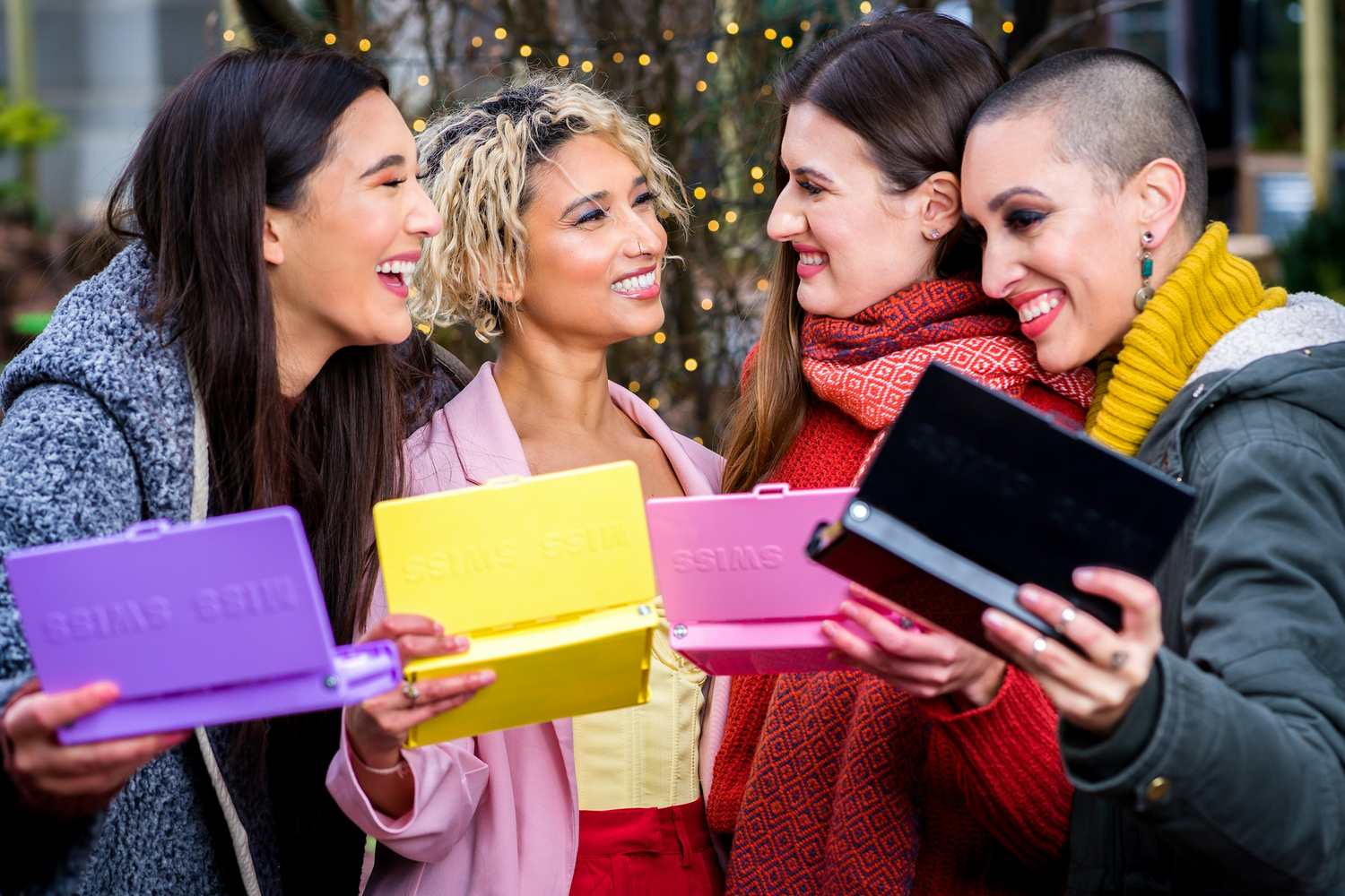 Four women holding colorful boxes with a blurred outdoor background