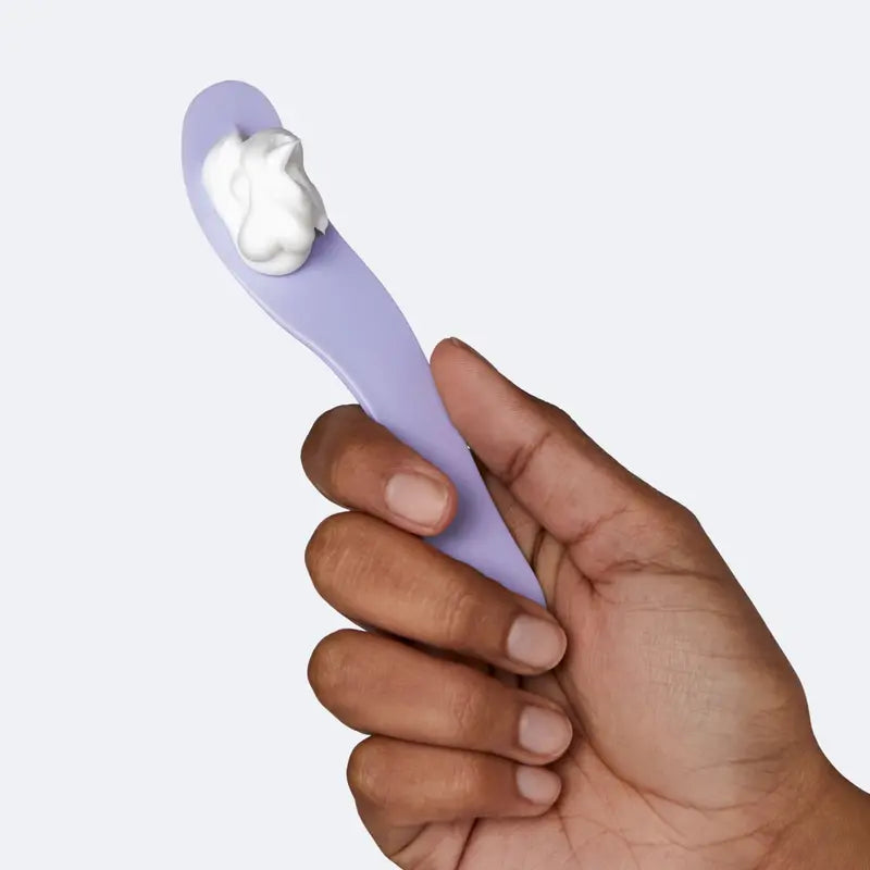 Hand holding a purple spoon with a scoop of white cream against a light gray background