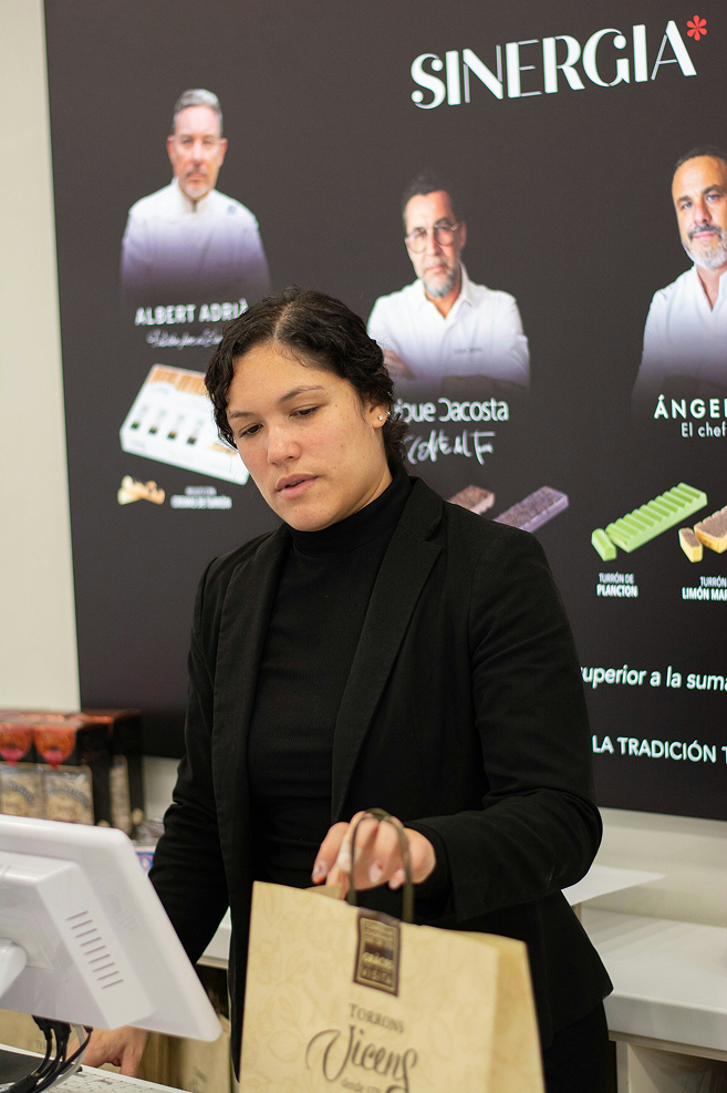 Woman in a black outfit holding a brown paper bag in front of a 'Sinergia' branded backdrop.