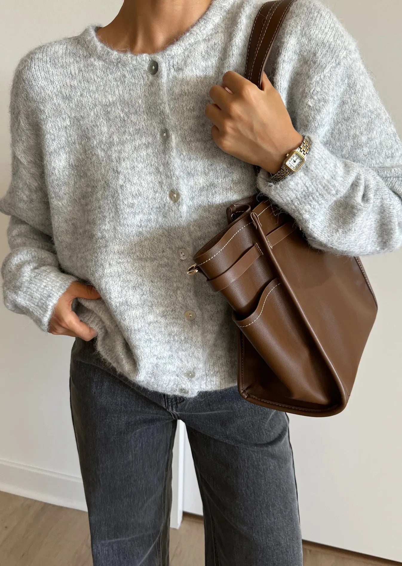 Person wearing a gray sweater and holding a brown leather bag indoors.