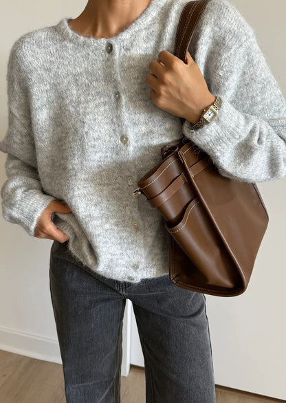 Person wearing a gray sweater and holding a brown leather bag indoors.